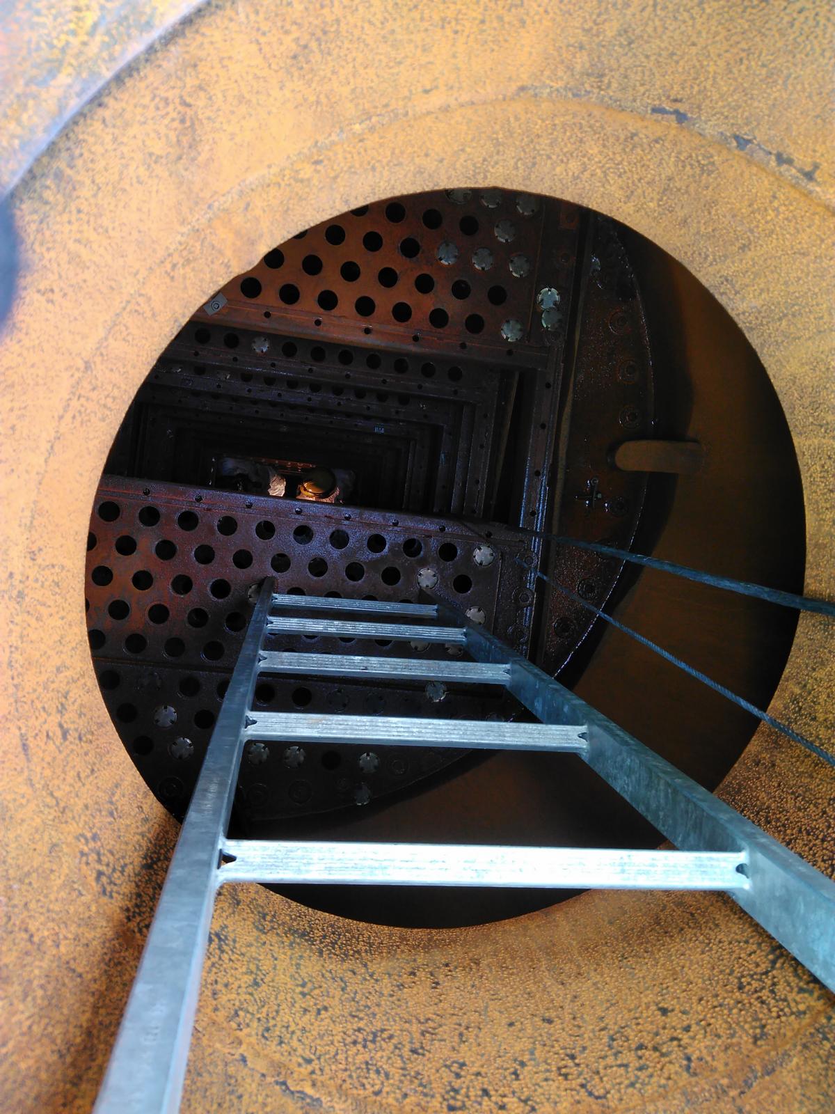 Blick hinunter in einen rostigen Metalltank, von dem eine Metallleiter nach unten führt.