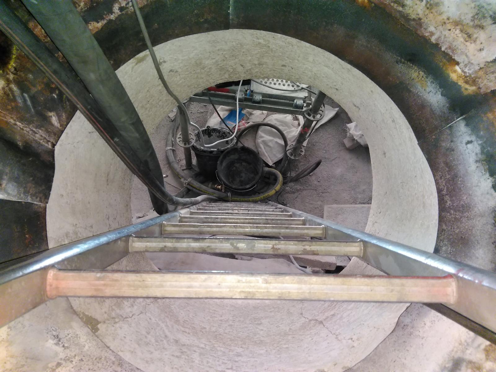 Blick von einer Leiter hinunter in einen zylindrischen Betonbunker, in dem sich am Boden Ausrüstung befindet.