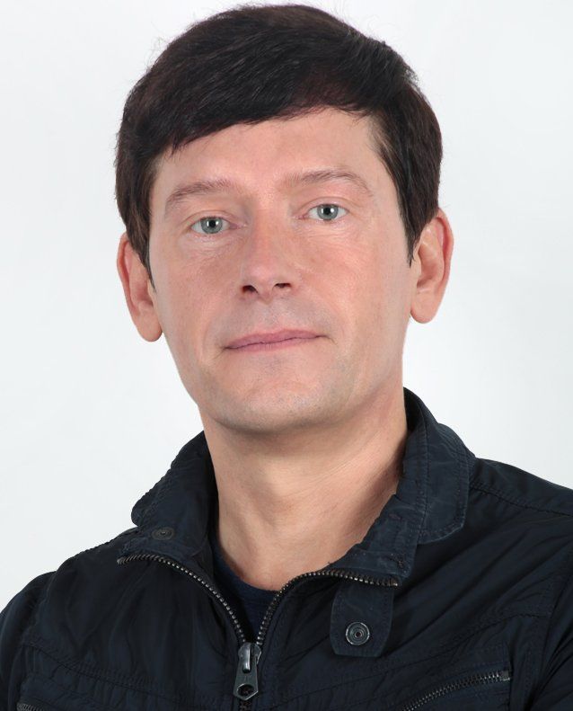 Ein Mann mit dunklen Haaren und blauen Augen, der eine dunkle Jacke trägt, blickt den Betrachter direkt an.