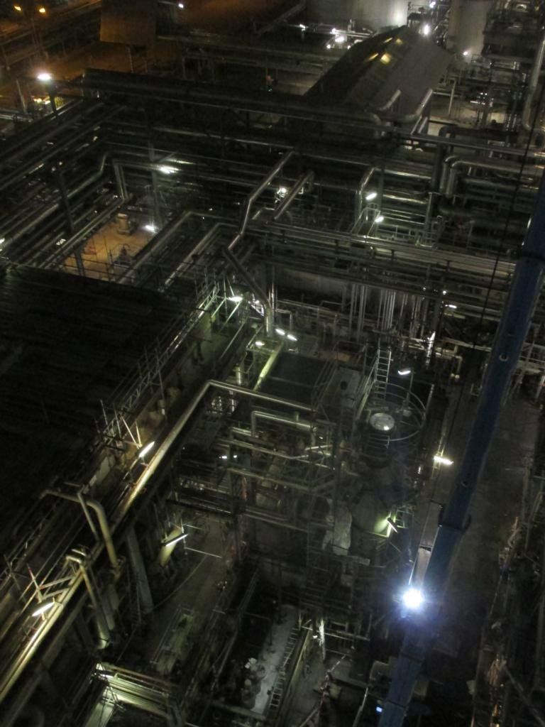 Komplexe Industrieanlage bei Nacht, beleuchtet von zahlreichen Lichtern und einem verzweigten Rohrleitungssystem.