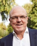 Ein Mann mit Brille und Glatze lächelt in einem Anzugjackett in natürlicher Umgebung.