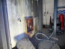 Rostige Metallzugangstür in einer Betonwand, im Hintergrund Schläuche und Geräte.