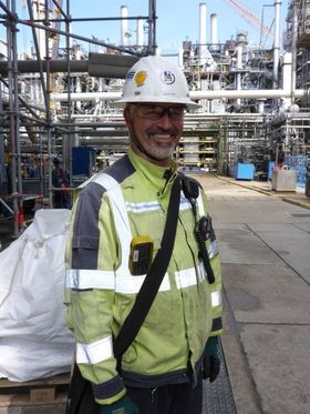 Ein Mann mit Schutzhelm und Sicherheitsweste lächelt vor einer Industrieanlage.