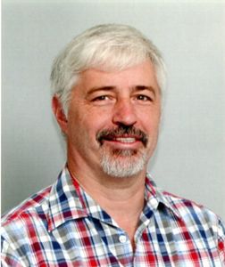 Mann mit grauem Haar und Bart, der ein kariertes Hemd trägt, lächelt in die Kamera.