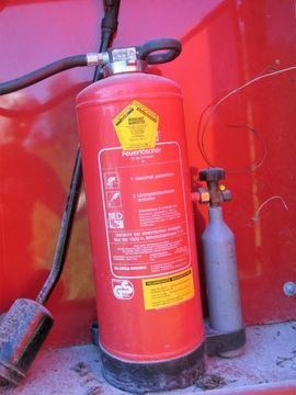 Ein roter Feuerlöscher ist in einem roten Gehäuse montiert. Inklusive Bedienungsanleitung und kleinem silbernen Zylinder.