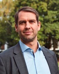 Ein Mann im Anzug und mit offenem Hemdkragen lächelt im Freien.