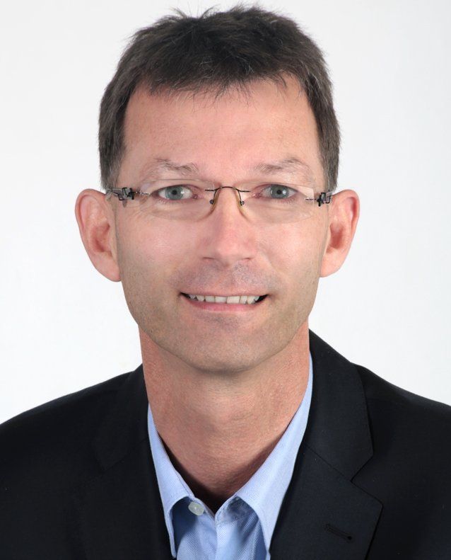 Mann mit Brille, der einen dunklen Anzug und ein hellblaues Hemd trägt, lächelt vor einem weißen Hintergrund.