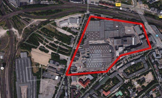 Luftaufnahme eines großen, rot umrandeten Industriekomplexes in der Nähe von Bahngleisen und städtischen Gebäuden.