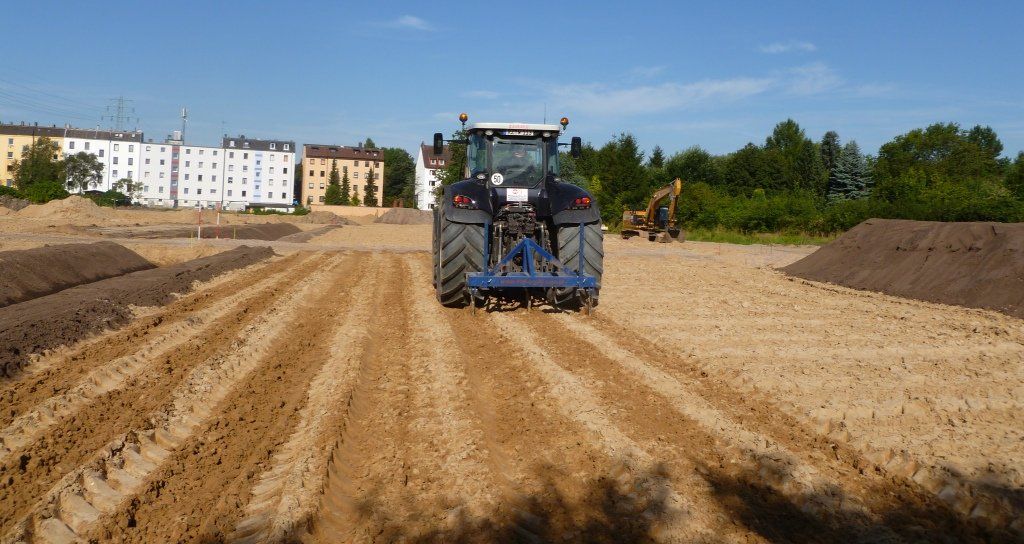 Ein Traktor bearbeitet ein Feld, im Hintergrund sind Gebäude unter blauem Himmel zu sehen.