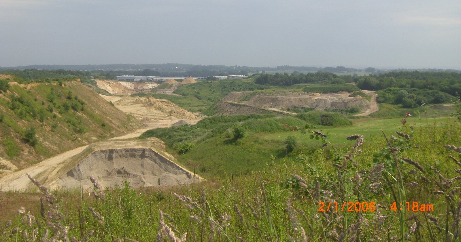 Landschaftsansicht eines Sandbruchs mit freiliegenden Erdwänden, grünem Gras und Bäumen unter einem bewölkten Himmel.