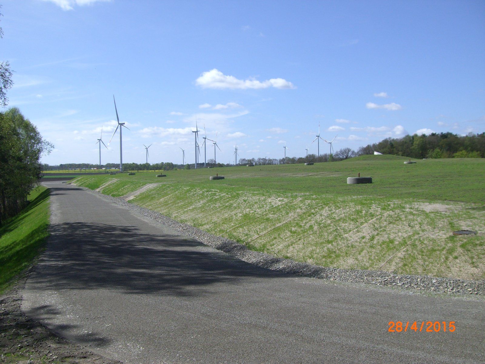 Ein Schotterweg führt zu Windkraftanlagen auf einem grasbewachsenen Feld unter blauem Himmel.