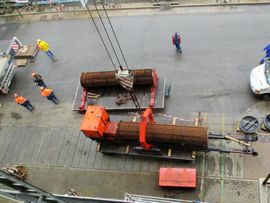 Ein Kran hebt große, zylindrische Gegenstände von einem Pritschenwagen; mehrere Arbeiter in Schutzkleidung.