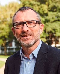 Mann mit Brille und Bart, der über einem blauen Hemd einen Blazer trägt, lächelt im Freien vor Bäumen.