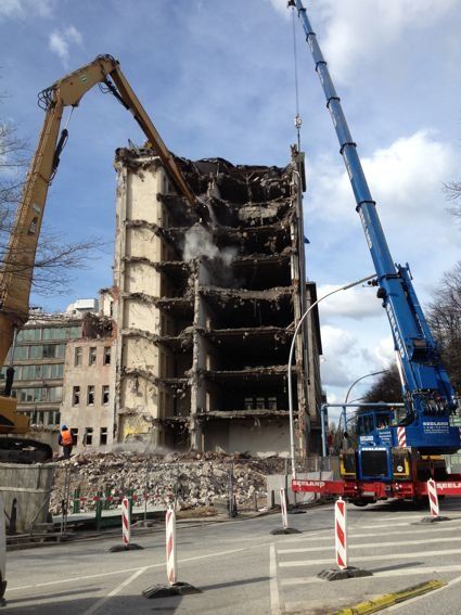Ein Gebäude wird von schwerem Gerät abgerissen, Rauch steigt auf, Straßenabsperrungen sind errichtet.