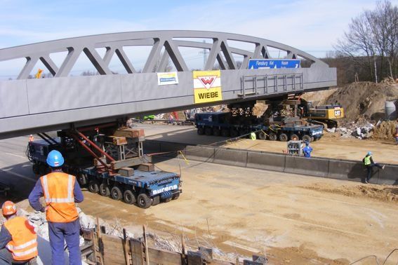 Eine große Stahlbrücke wird von mehreren Schwerlastwagen und Bauarbeitern über eine Fahrbahn bewegt.