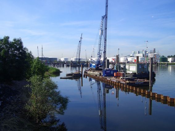 Baumaschinen auf einem Fluss, im Hintergrund Öltanks unter blauem Himmel.