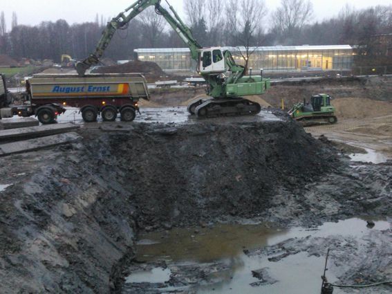 Ein Bagger belädt einen Muldenkipper mit Erde auf einer Baustelle; im Hintergrund ist ein Gebäude zu sehen.