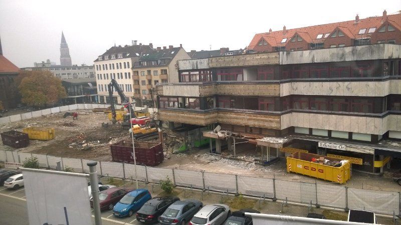 Baustelle mit einem abzureißenden Gebäude; gelbe Müllcontainer und schwere Baumaschinen sind zu sehen.