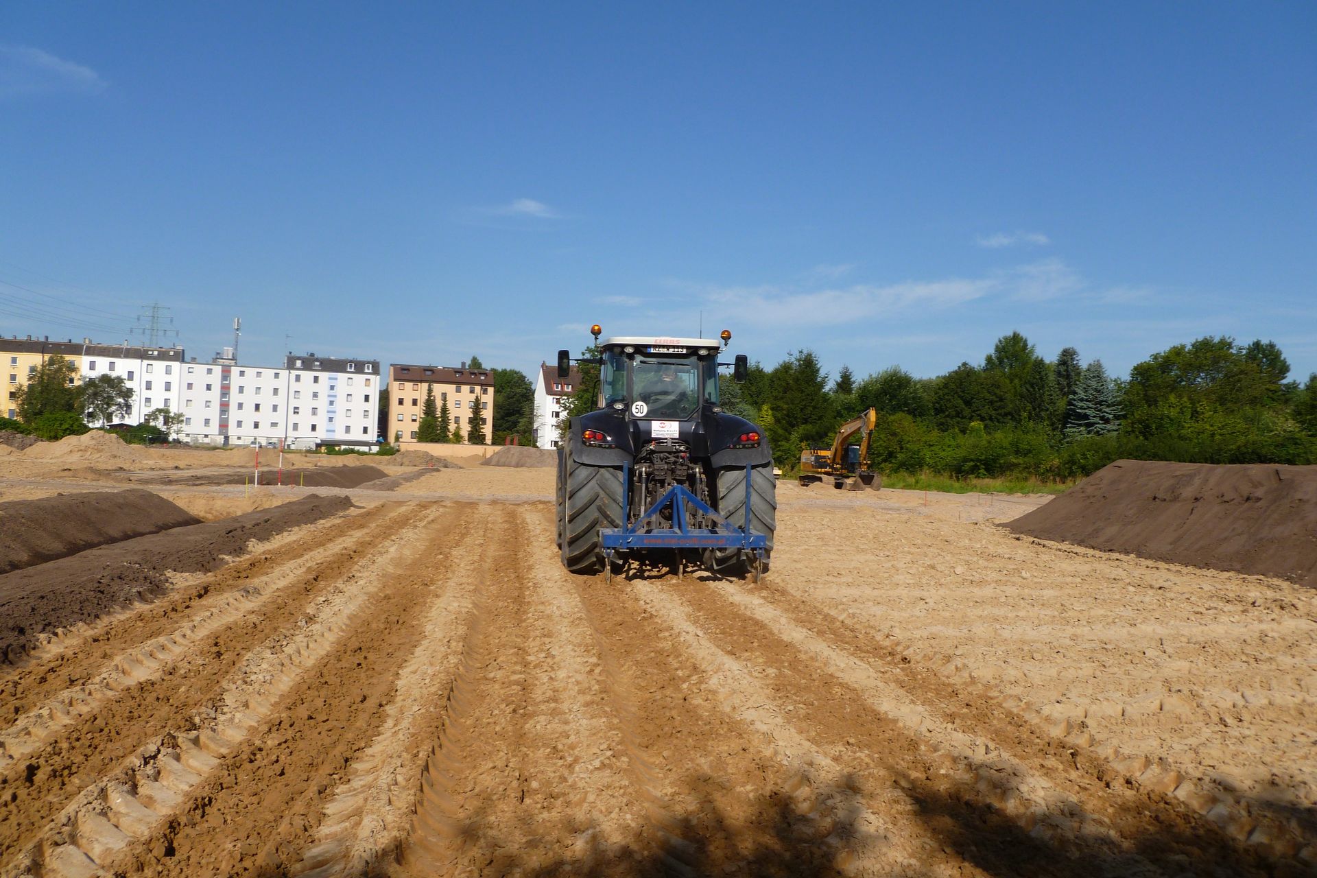 Ein Traktor pflügt ein Feld, brauner Boden, blauer Himmel, Gebäude im Hintergrund.