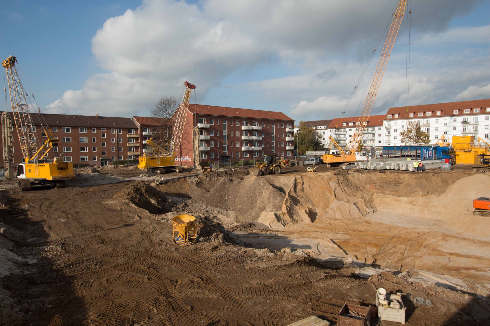 Baustelle mit Gebäuden im Hintergrund; Erdbewegungsmaschinen und Kräne arbeiten unter bewölktem Himmel an den Aushubarbeiten.