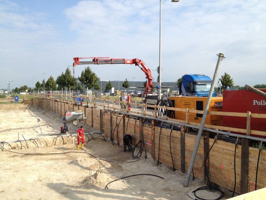 Baustelle mit Stützmauer, Arbeitern und Maschinen, darunter eine Betonpumpe.