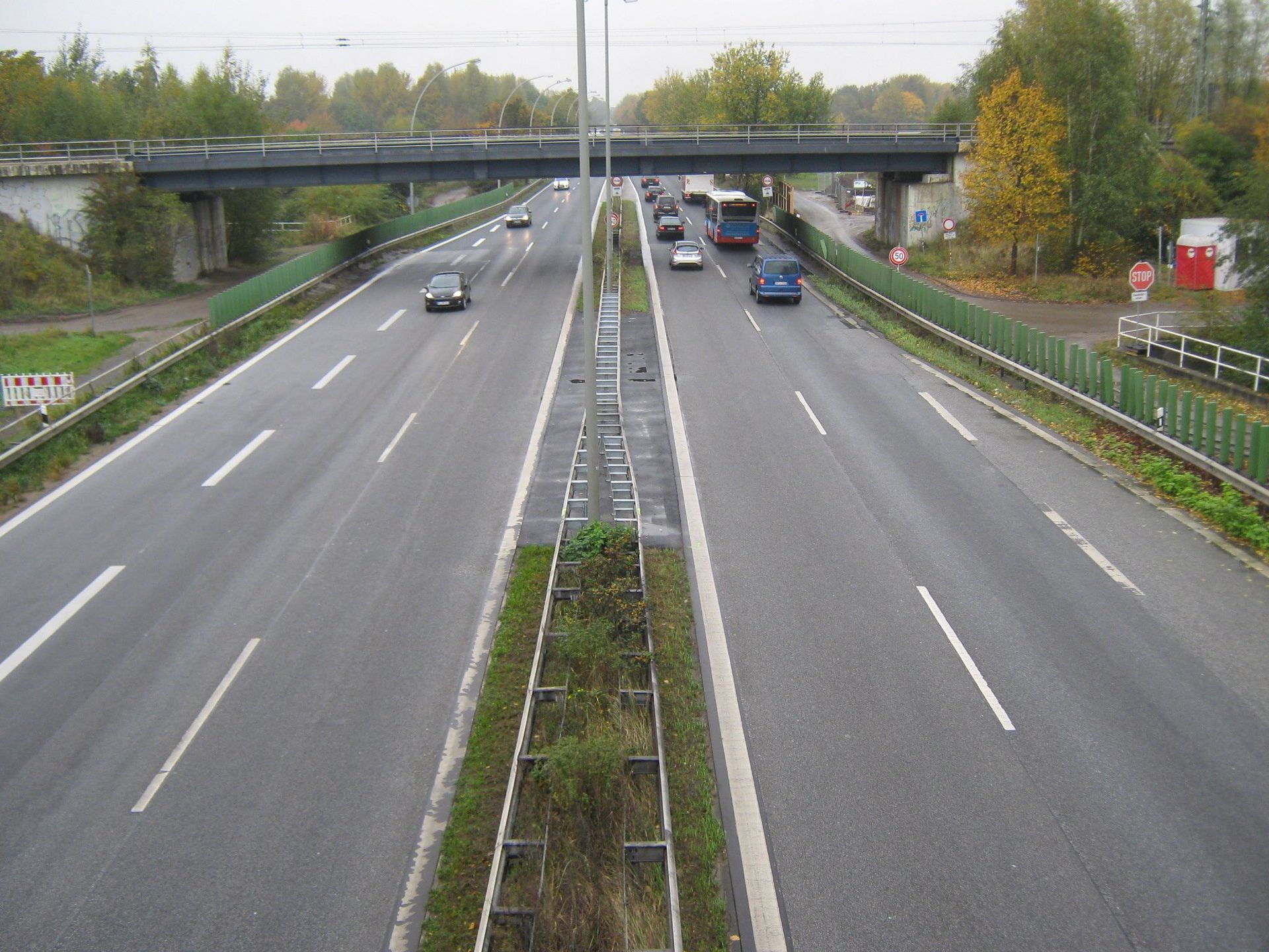 Autobahn mit mehreren Fahrspuren, Fahrzeugen, Überführung und grünen Leitplanken. Graue Straße, bewölkter Tag.