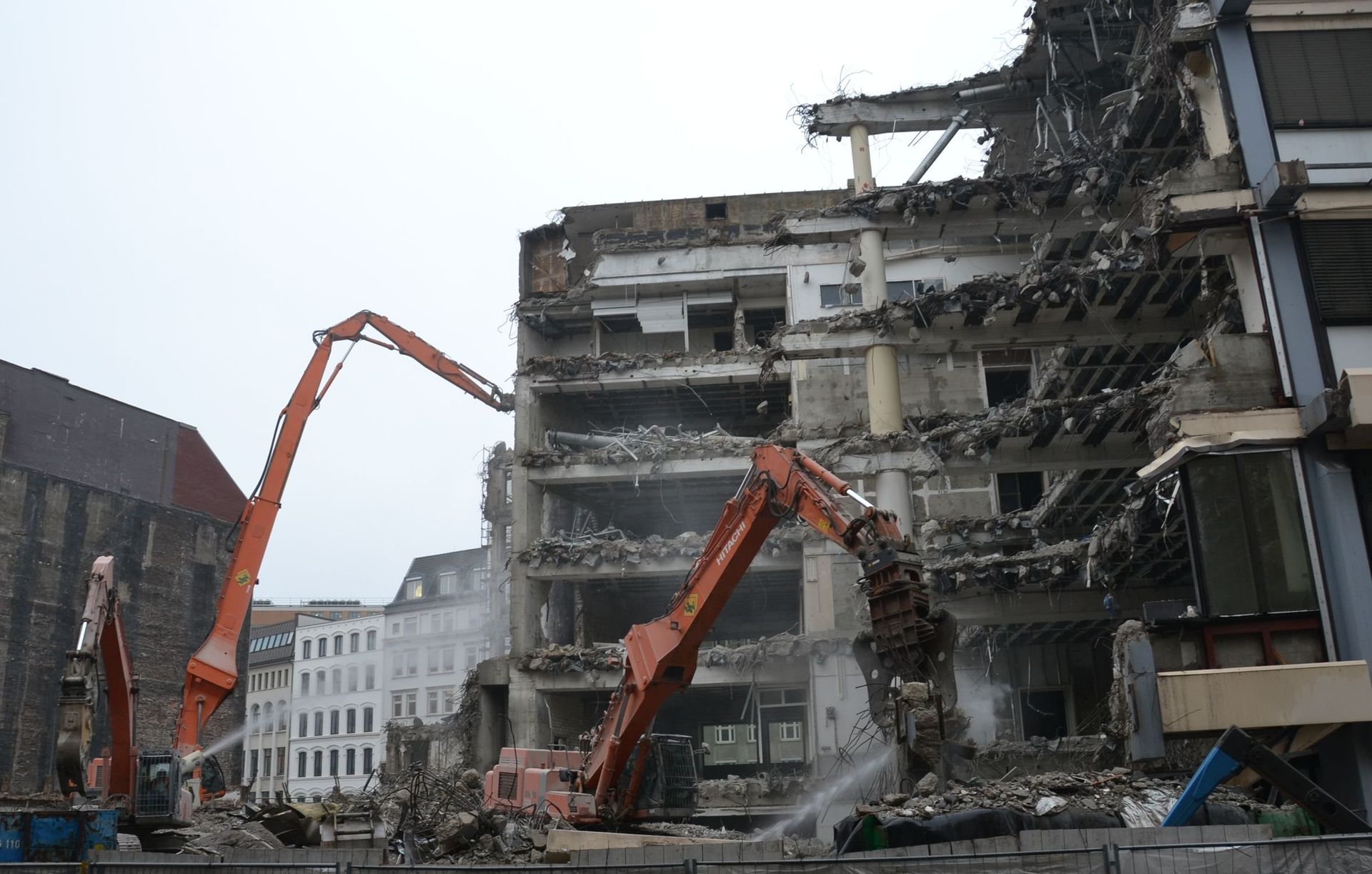 Zwei orangefarbene Bagger reißen ein mehrstöckiges Gebäude ab, Trümmer liegen überall herum.