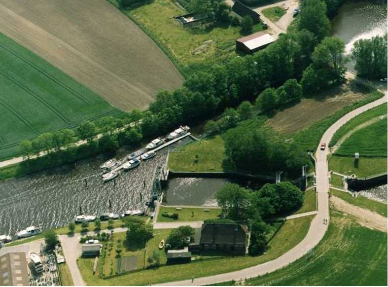 Luftaufnahme einer Kanalschleuse mit Booten, umgeben von grünen Feldern, Bäumen und einer gewundenen Straße.