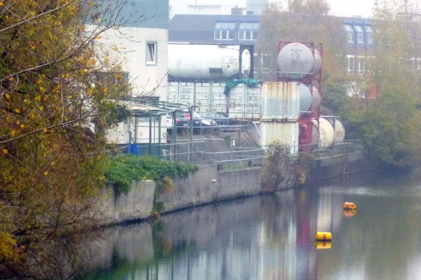 Flussuferblick mit Industrietanks und Gebäuden am Ufer; Herbstlaub.