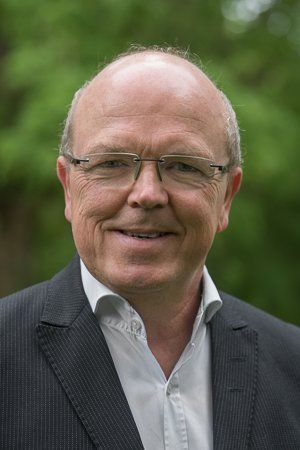 Mann mit Brille, der ein Sakko und ein weißes Hemd trägt, lächelt vor einem verschwommenen grünen Hintergrund.