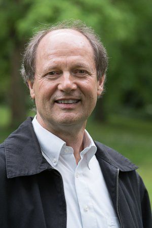 Ein Mann lächelt im Freien vor grünem Hintergrund. Er trägt eine schwarze Jacke und ein weißes Hemd.