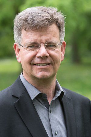 Mann mit Brille, dunklem Anzug und grauem Hemd lächelt, im Freien vor verschwommenem Grün.
