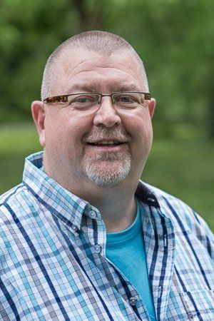 Mann mit Brille und Spitzbart lächelt, trägt ein blau kariertes Hemd, im Freien vor verschwommenem grünem Hintergrund.