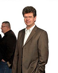 Mann im braunen Anzug, weißem Hemd und Brille, lächelnd, die Hand in der Tasche, steht.