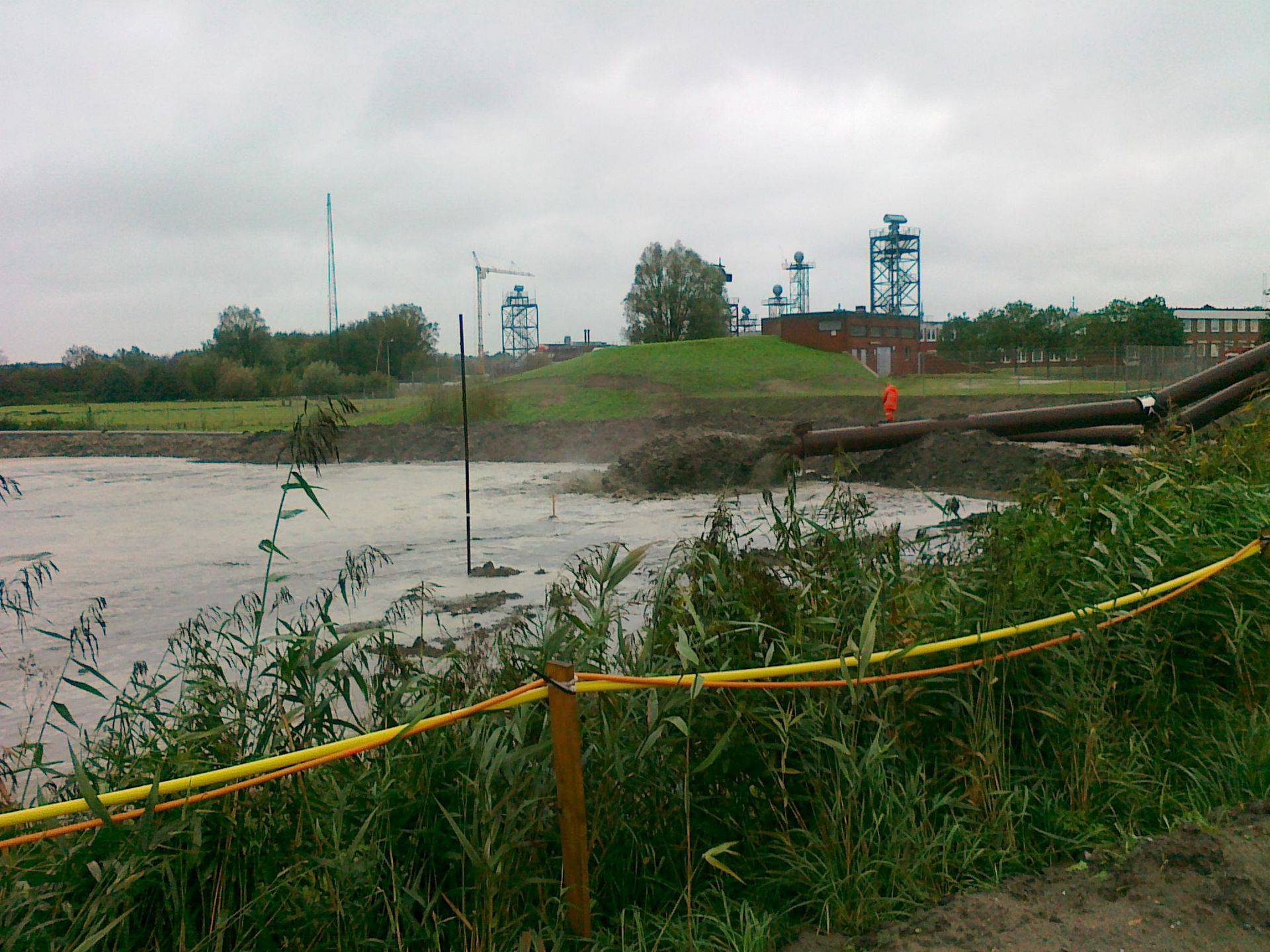 Trübes Wasser, Industrieanlage im Hintergrund; gelbe Barriere im Vordergrund. Bewölkter Himmel.