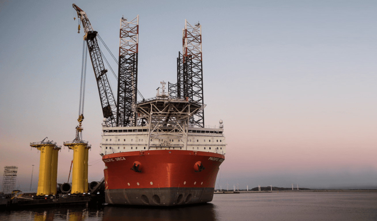Großes rotes Schiff mit Kran, gelbe Strukturen und hohe Metallstützkonstruktionen in einem Hafen in der Abenddämmerung.