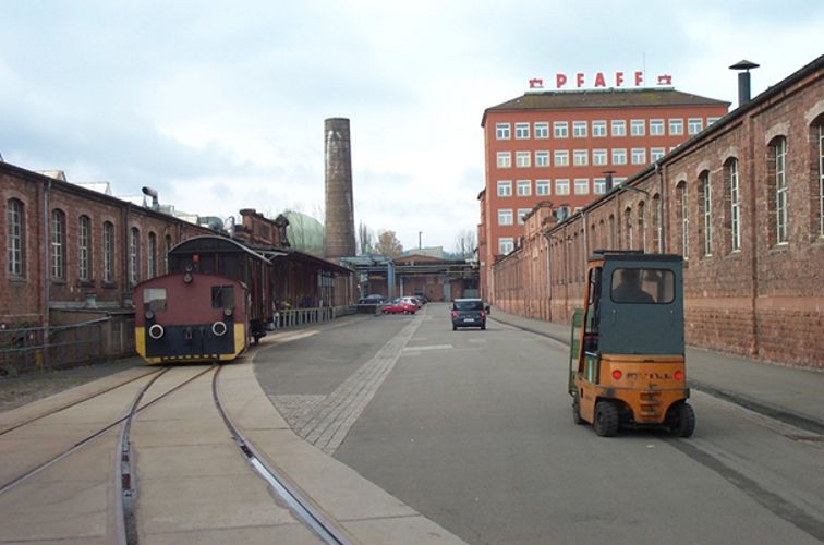 Industrieszene mit einem Zug auf Gleisen, einem Gabelstapler, Backsteingebäuden und einem „PFAFF“-Schild.