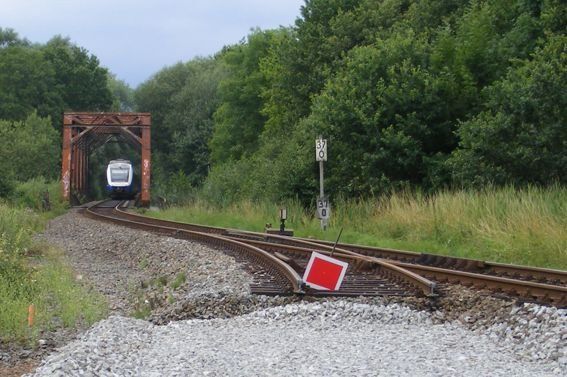 Ein Zug nähert sich einem roten Signal und verlässt eine braune Brücke. Im Hintergrund sind Gleise, Grün und Himmel zu sehen.