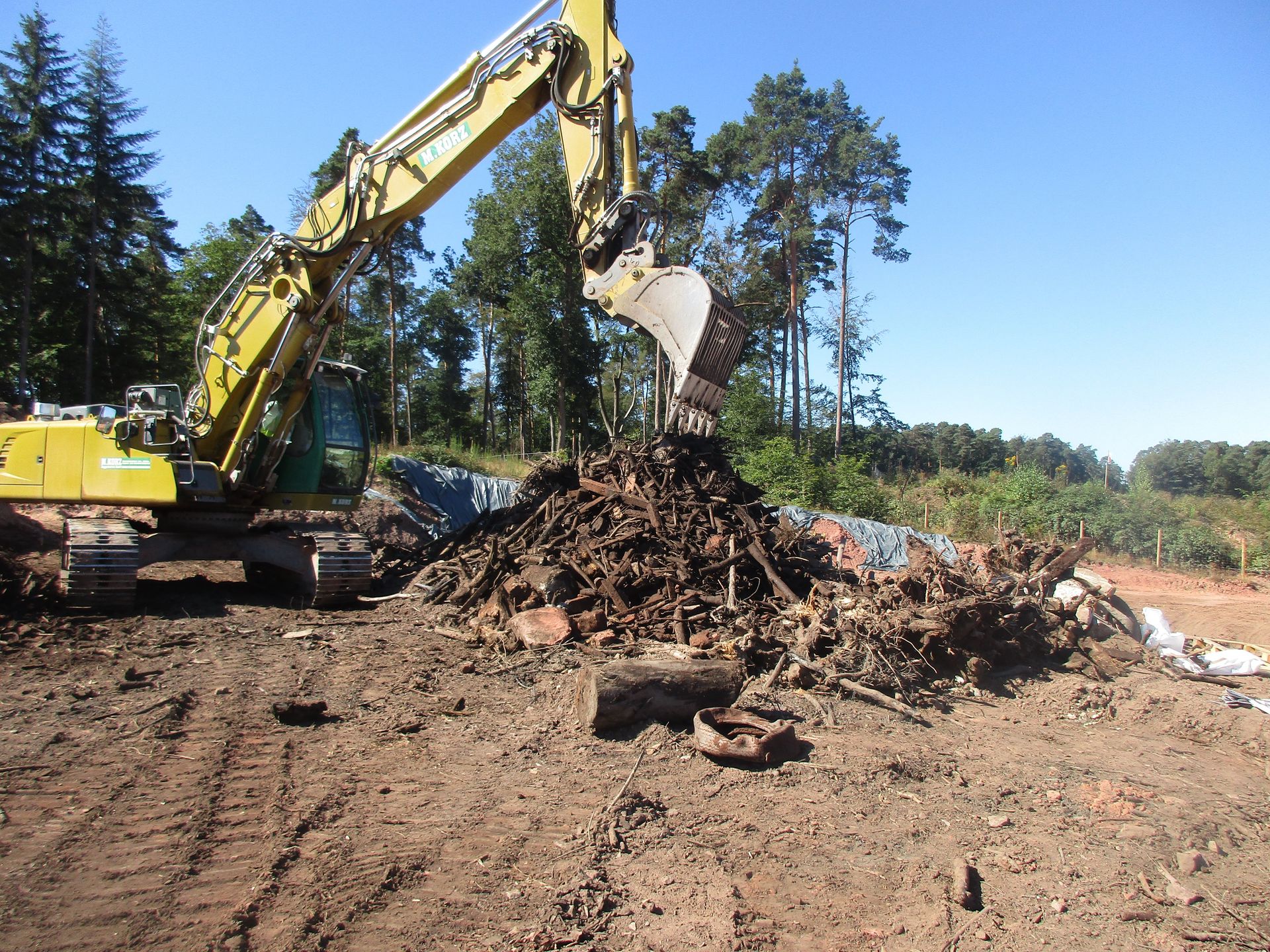Ein gelber Bagger gräbt Schutt auf einer unbefestigten Fläche aus; im Hintergrund ist ein Wald zu sehen.