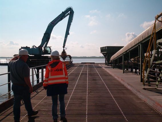 Zwei Personen in Schutzausrüstung inspizieren unter blauem Himmel ein Dock mit Industriemaschinen.
