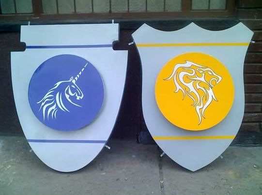Dos letreros con forma de escudo y logotipos de unicornios, uno azul y otro amarillo, apoyados contra una pared.