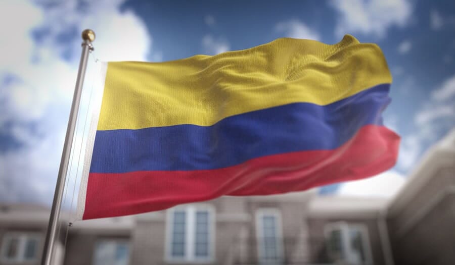 Bandera colombiana ondeando en un mástil contra un edificio borroso de fondo.