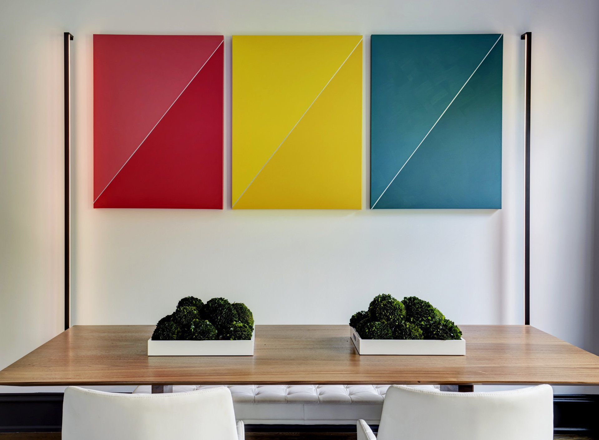 A dining room table with three colorful paintings on the wall above it