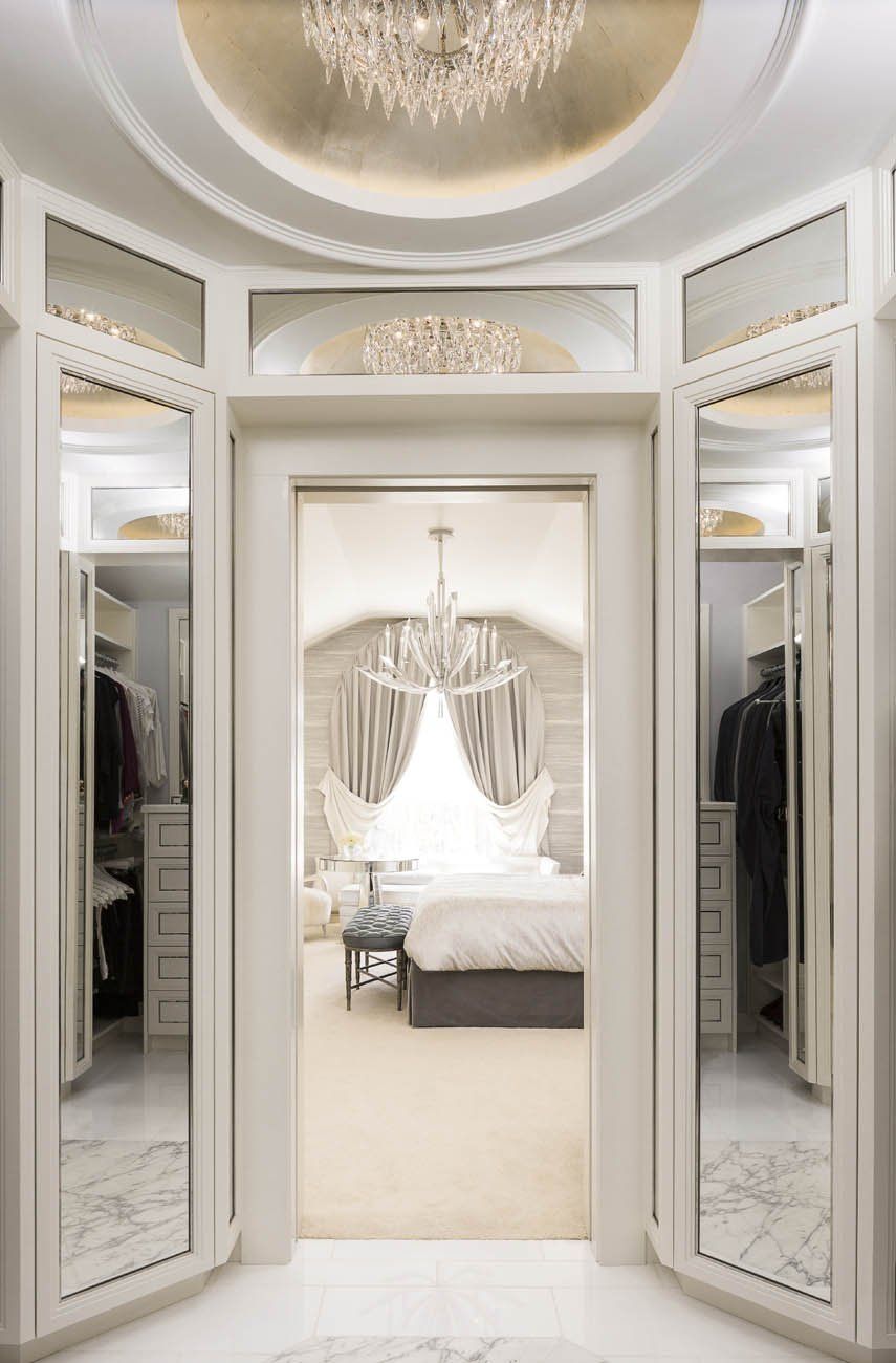 A walk in closet with a chandelier hanging from the ceiling and a bedroom in the background.