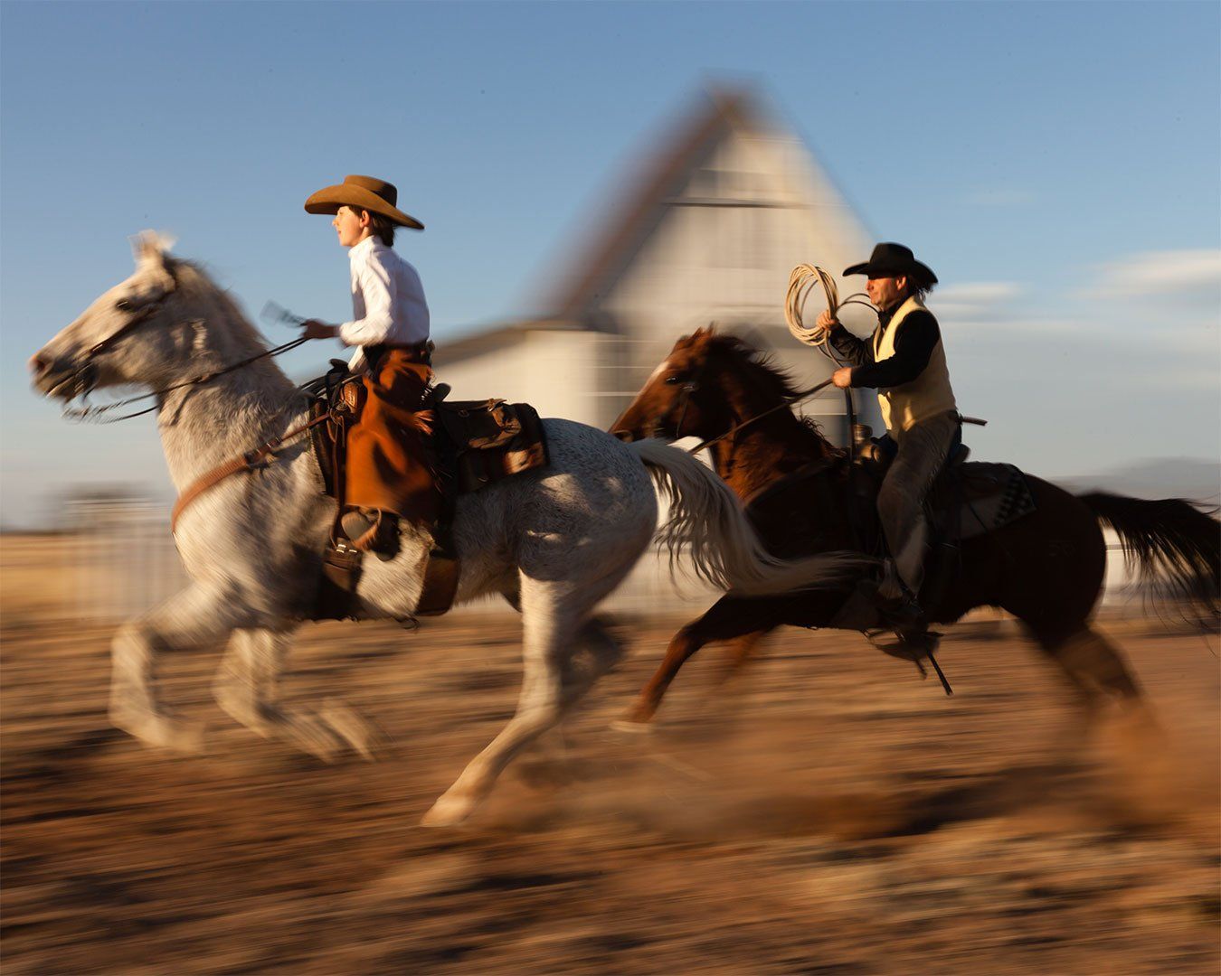 Two cowboys on horseback, one with a lasso, riding across a dry field near a white barn.
