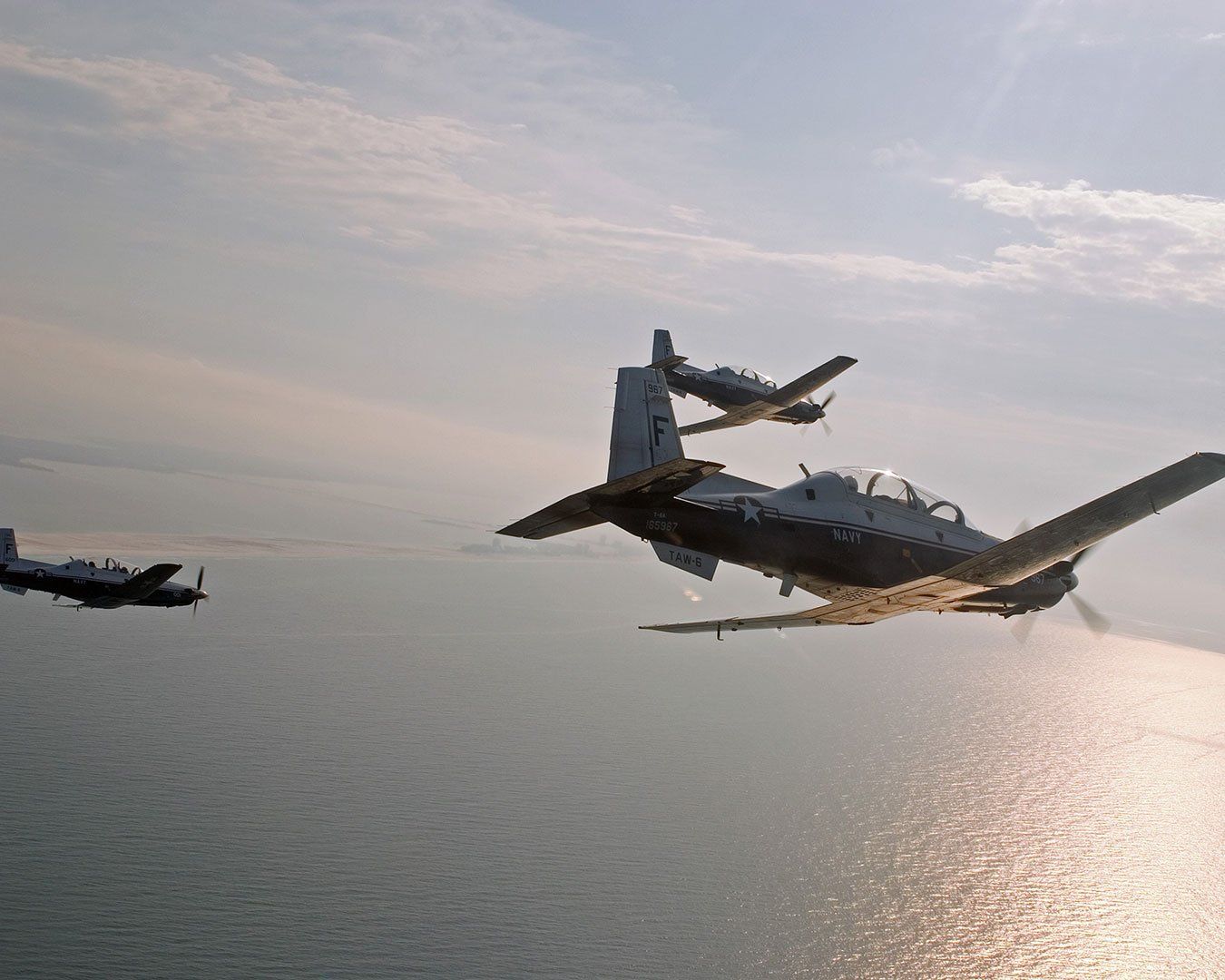 Three military training aircraft in formation over a body of water, against a hazy sky.