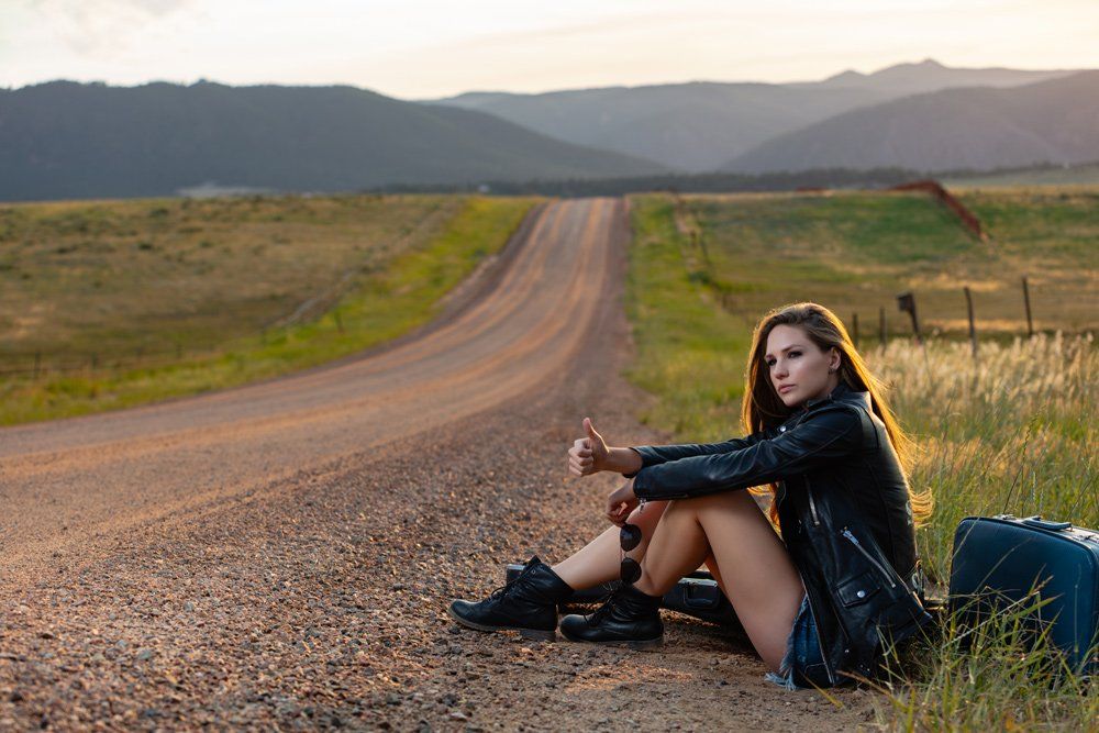 Woman hitchhiking on a rural road, thumb up, sitting next to a suitcase. Golden sunlight, mountains in the background.
