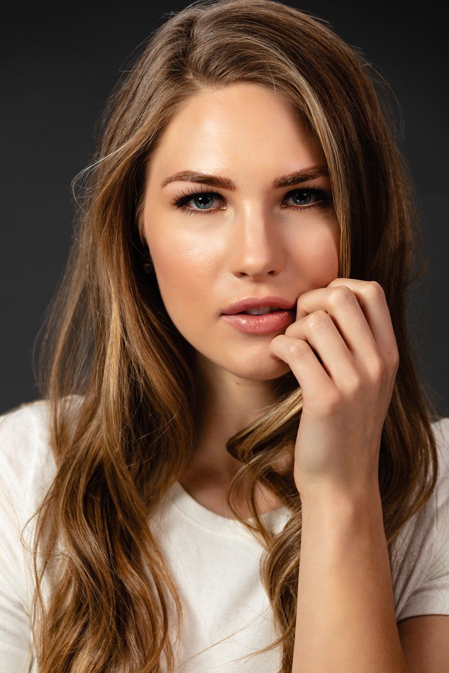 Woman with long, wavy hair, in a white t-shirt, looking at the camera. She's touching her face, with a soft expression.