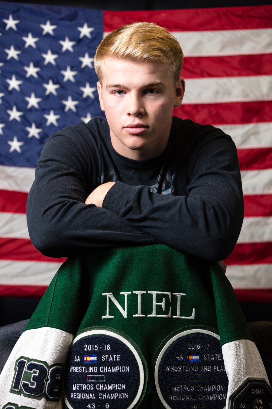 Young man in front of American flag, arms crossed. Green letterman jacket draped over his lap. Blonde hair.