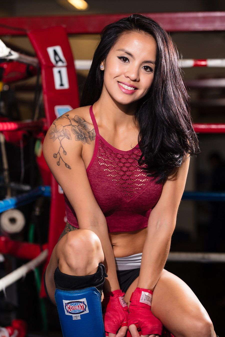 Woman in boxing ring, wearing red top, wrapped hands. Tattoo on shoulder, smiling.